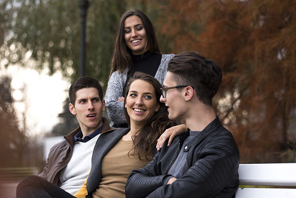Four young friends, two girls and two guys, in casual clothes sitting in a park on a white bench in autumn and smiling while looking at one of guys.